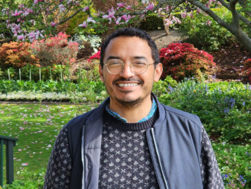 Man in a blue jacket ,patterned jumper and glasses standing in a park