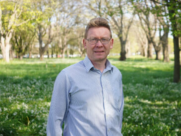 Man in light coloured shirt standing in a park on a sunny day
