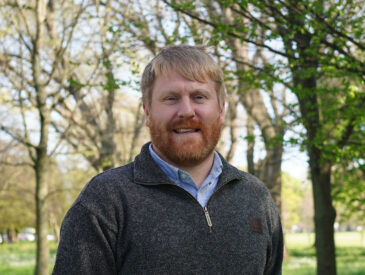 Man with blonde hair and beard wearing a grey jumper standing in a park with trees in the background