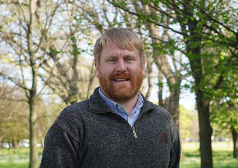 Man with blonde hair and beard wearing a grey jumper standing in a park with trees in the background