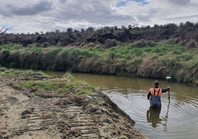 View of man dressed in orange and waterproof gear waist deep in the river surrounded by undergrowth on an overcast day.