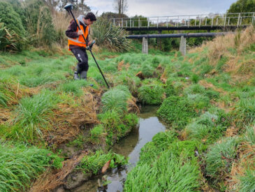 Man dressed in orange and grey holding surveying equipment on the side of a river