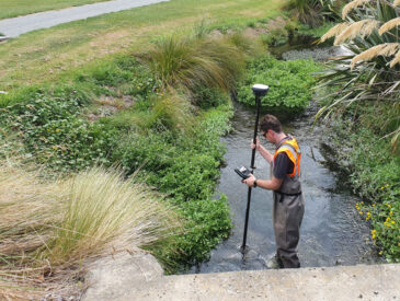 Man dressed in orange and grey waterproof clothing holding surveying equipment knee-deep in a river
