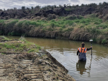 View of man dressed in orange and waterproof gear waist deep in the river surrounded by undergrowth on an overcast day.