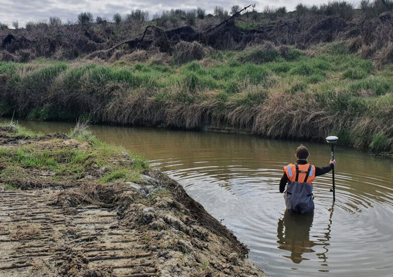 View of man dressed in orange and waterproof gear waist deep in the river surrounded by undergrowth on an overcast day.
