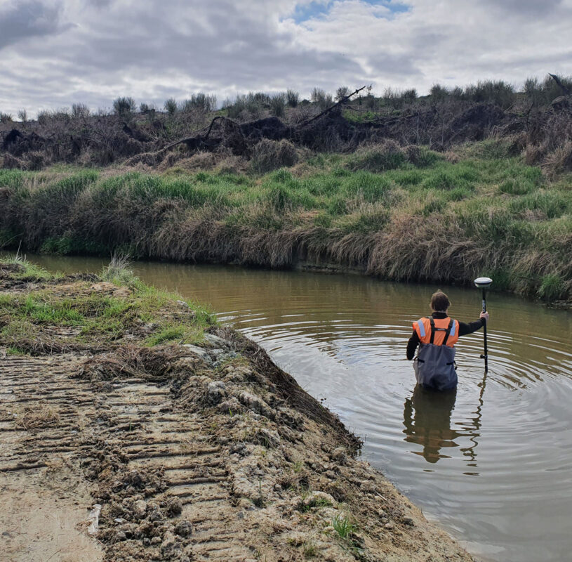 View of man dressed in orange and waterproof gear waist deep in the river surrounded by undergrowth on an overcast day.