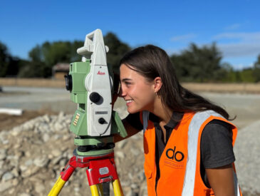 Women dressed in high visibility vest looking into survey equipment outside with blue sky in the background