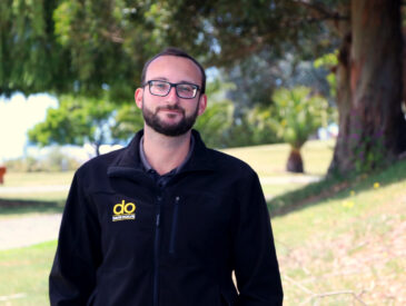 Man with dark hair, wearing a dark DO branded top and glasses in a park in the background