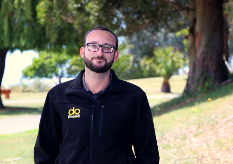 Man with dark hair, wearing a dark DO branded top and glasses in a park in the background